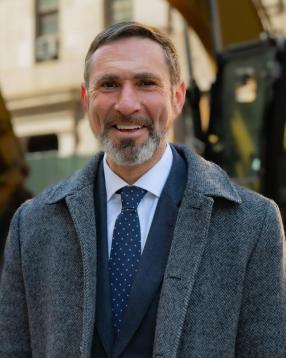 Jamie Torres-Springer from the shoulders up wearing a gray overcoat, blue suit, white shirt, and blue tie, with a construction site in the background