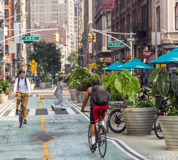 People ride bicycles along a two-way green bike lane along a pedestrian plaza in Manhattan on a sunny day.
