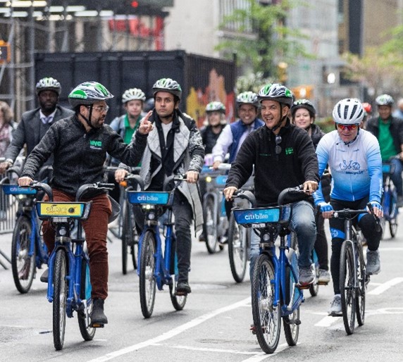 A group of people wearing bicycle helmets ride blue CitiBikes along a car-free avenue in Manhattan.